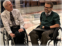 On an elaborate marble floor, two men in business dress, in manual wheelchairs, sit laughing.