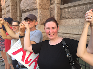 Erin smiles at the camera from a line of people with upraised, joined hands. The stone façade of the Texas Capitol is right behind them. 