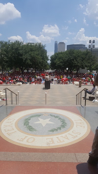 From behind, a man at a podium holds an arm in the air as he addresses a crowd. They are outside, and in the foreground, is the seal of the State of Texas in marble flooring.