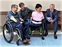 On a wooden bench, a man in a blue suit flips through a booklet. Next to him, with their attention on the booklet are a smiling young man with no arms or legs in a wheelchair, a pensive young man sitting on the other side of the bench, and a man in a wheelchair with glasses.