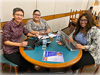 Three women sitting at a teal cafeteria table look up from their laptops to smile at the camera.