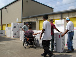 Outside a warehouse with stacks of white boxes, a woman sitting in a wheelchair with her back to camera looks through papers, while several men move boxes.