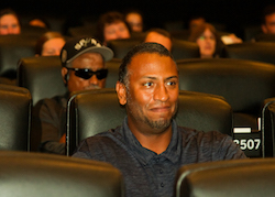A man sitting among rows of theater seats looks up and off camera and holds back a big smile