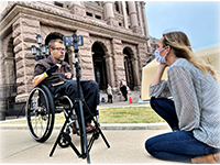 Shot from a low angle, a woman kneels behind a phone on a tripod, recording a man gesturing from a manual wheelchair. The Texas Capitol building is in the background.