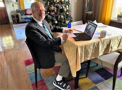 A man in a tie and jacket (and sandals) sits at a square dining with a laptop open in front of him. A Christmas tree is visible in the background.