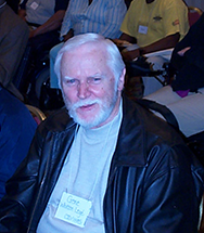 A man with white hair and beard smiles up at the camera from a folding chair among a big group seated people.