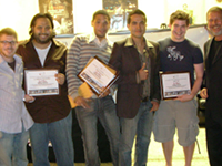 Six men, five college-age and one older, stand in a line holding up certificates, smiling broadly at the camera.