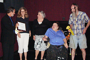 A man hands a plaque to a smiling woman. Next to her are three people smiling and looking on, one sitting in a power chair, another holding a yellow flower.