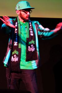 A man in a green and black hat, scarf, and jacket holds his hands out in front of him, blurred in motion.