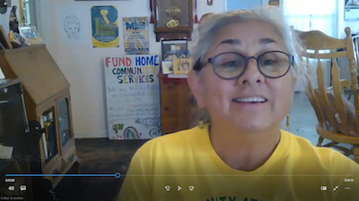 Cathy smiles from her Zoom window. She's wearing a bright yellow shirt and glasses, and protest signs and posters can be seen in the background.