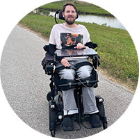 A smling, bearded man sits in a powerchair, resting one hand on an attached clear tray, He is on a paved path, running through a grassy field with a stream.