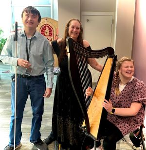 Three smiling people surround a small harp. A woman in the center puts her hand on the shoulder of a young man with a white cane. Her other hand is on the shoulder of a young seated woman with her hands at the harpstrings.