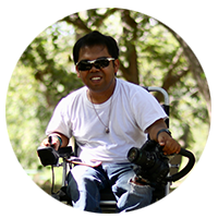 In a shady wooded area, a man with sunglasses sitting in a power chair smiles at the camera.