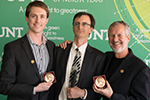 Three men in suits smile broadly at the camera. Two hold bronze, circular awards and the other grips a white cane.