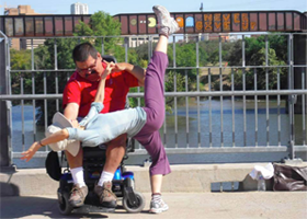 On a bridge with a train rolling by are a pair of dancers. One is in a power chair, and he holds up one hand of the other, who leans back across his knee and lifts one leg high in the air.