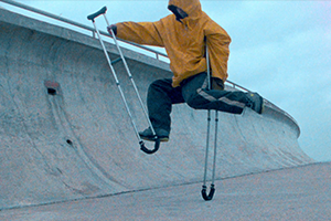A figure in a puffy yellow jacket that obscures his face dances on crutches in front of a curving concrete wall. He is suspended on one crutch, one arm and leg wrapped around it, while his other crutch, arm, and leg unwind in front of him.