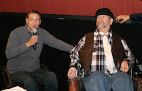 A man seated in a wheelchair speaks into a microphone. He has his hand resting on the powerchair of another man, who listens with an uneasy expression.
