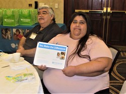 Anita, sitting at a banquet table, holds up a certificate to the camera. A man sitting next to her appears to be listening to something off camera.