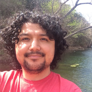 With a river and trees in the background, a man in a red tshirt smiles at the camera.