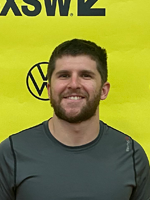 Evan, a bearded man in a gray t-shirt, smiles at the camera, in front of a yellow SXSW banner