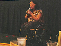 A woman sitting in a wheelchair holds a microphone close to her face with both hands.