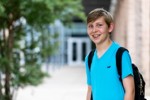 Standing in 3/4 profile to the camera is a smiling young man with braces and a backpack