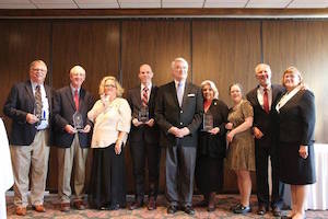A group of men and women of mixed ages and business attire stand in a line and pose for the camera, most are holding glass plaques.