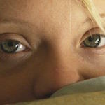 Close up on a young woman's face with strands of blonde hair and green eyes. She has green blankets pulled up to her nose and stares directly at the camera.