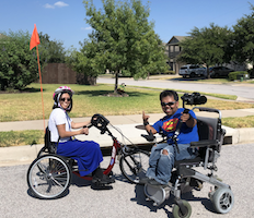 On a bright residential street corner, woman in a sit-down bike and a man in a power chair with video camera rig smile excitedly at the camera. He's giving the shaka sign.