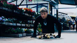 Shot from a low angle, a young man with no legs and a determined expression leans forward on a skateboard as he propels himself with his hands.