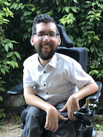 Surrounded by leafy bushes, a smiling man with a beard and glasses leans forward to the camera in his power chair.
