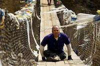 Wooden bridge with with mostly white strips of cloth tied to netted handrails. Shot from head-on, a man makes his way across the bridge on his hands and bottom, with his legs folded under him.