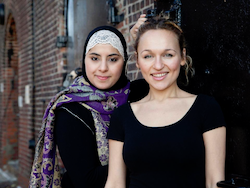 two women stand close together against a brick wall, smiling at the camera. One wears a hijab