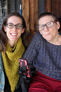 Laura and Susie posing for a portrait on the front porch of CTD. Laura wears black glasses and a mustard colored scarf. Susie also wears black glasses and has on a grey cable knit sweater. Her power chair armrest is visible, holding her hot pink phone. They're both smiling, Susie looks like she might be up to something.