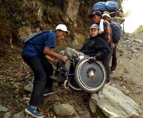 On an upward sloping dirt trail, two men hoist a third in a wheelchair over some rocks.