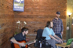 From the side, a trio of male musicians are set up in a corner with wood paneling. One sits and plays a guitar, another sits in a power chair with his hands on a laptop, and a third stands to play a snare drum.