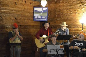 A group of five older men play instruments, sing, and look at sheet music in a wood-paneled room. 