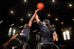 two wheelchair basketball players at tip off in a dark arena, one facing toward the camera, the other with his back to the camera, each reaching for the ball in the air.