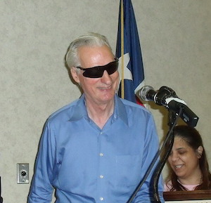 A tall smiling man with white hair and dark glasses approaches a podium with a microphone.