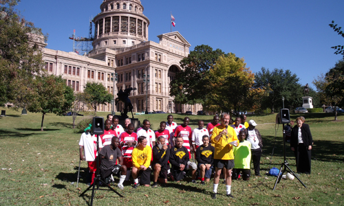 group of soccer players pose together while a man speaks into a microphone, with Texas state capitol in the background